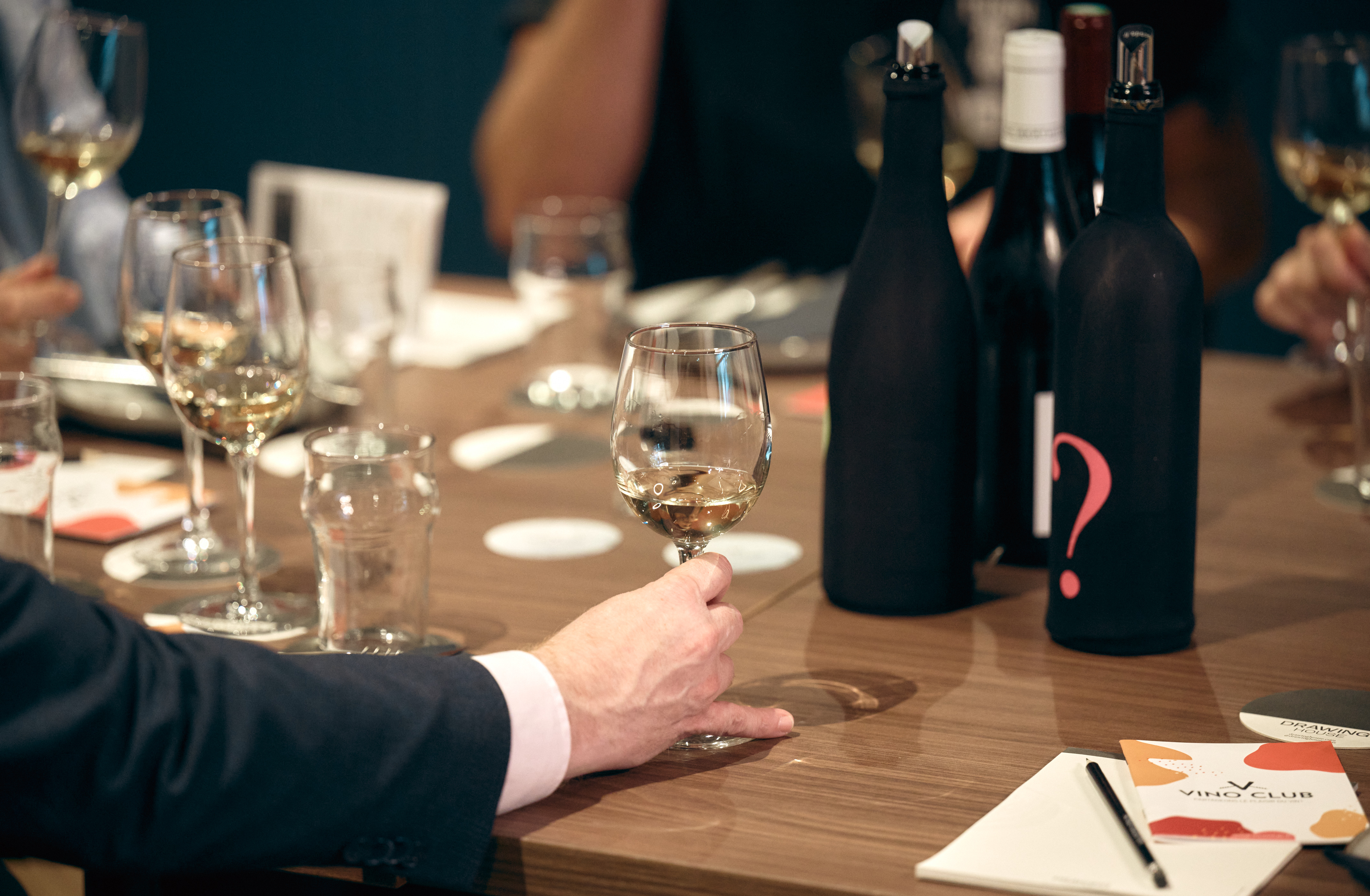 Dégustation de vin lors d’un atelier œnologique avec des bouteilles mystères. Un participant en costume tient un verre de vin blanc, tandis que d'autres convives analysent les arômes et saveurs. L’événement est organisé par Vino Club dans un cadre convivial et raffiné.
